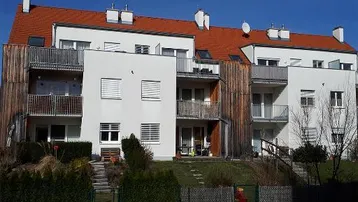 Modernes Mehrfamilienhaus mit weißer Fassade, roten Ziegeldächern und mehreren Balkonen, umgeben von einem gepflegten Garten.
