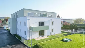 Modernes Mehrfamilienhaus mit weißer und grüner Fassade, umgeben von einer Rasenfläche und einem kleinen Gartenbereich.