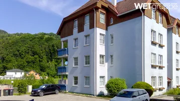 Mehrstöckiges Apartmentgebäude mit hellblauer Fassade, Holzelementen und Balkonen, umgeben von grüner Natur.