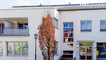 Moderne Gebäudefassade mit weißem Anstrich und sichtbarem Balkon, umgeben von Herbstbäumen.