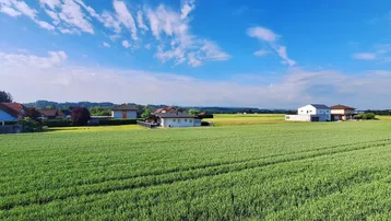 Panoramablick über weite grüne Felder, ländliche Häuser und ferne Berge unter blauem Himmel.