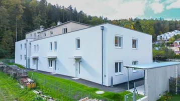 Modernes Mehrfamilienhaus mit weißer Fassade in grüner Hanglage, umgeben von Wald.