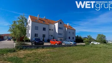 Mehrfamilienhaus mit weißer Fassade und rotem Satteldach, umgeben von Grünflächen und Parkplätzen.