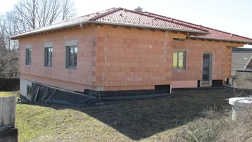 Haus im Rohbauzustand mit Ziegelwänden und Dachziegeln, umgeben von einer Baustelle.
