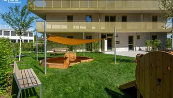 Grüner Gemeinschaftsgarten mit Sandkasten, Sitzbänken und Sonnenschutz vor einem modernen Wohngebäude.