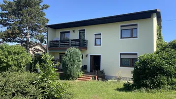 Einfamilienhaus mit hellbeiger Fassade, Balkon und großem Gartenbereich unter blauem Himmel.