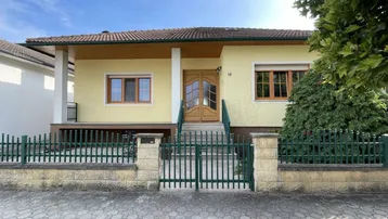 Frontansicht eines Einfamilienhauses mit gelber Fassade und grünem Zaun unter blauem Himmel.