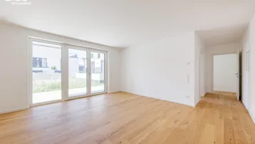 Helles, leeres Zimmer mit Holzboden und großen Fenstern, die viel Tageslicht hereinlassen.