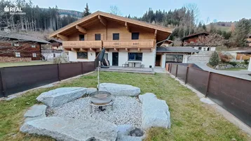 Traditionelles Holzchalet mit weißer Fassade, umgeben von einem Garten mit Feuerstelle und Bergblick.