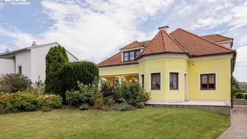 Charmantes gelbes Haus mit rotem Ziegeldach, umgeben von einem gepflegten Vorgarten und blühenden Sträuchern.