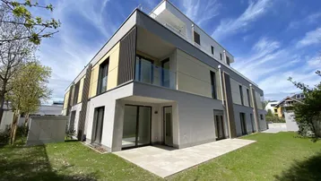 Modernes Mehrfamilienhaus mit gepflegtem Garten, großzügiger Terrasse und Balkonen unter blauem Himmel.