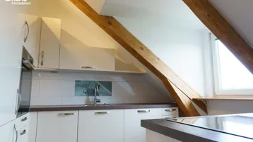 Moderne Einbauküche im Dachgeschoss mit weißen Schränken, dunkler Arbeitsplatte und Holzbalken.