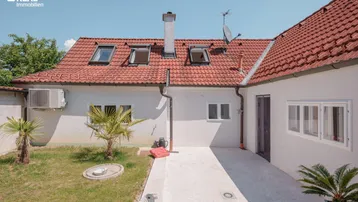 Hausrückseite mit kleiner Rasenfläche, gepflasterter Terrasse und Dachfenstern unter blauem Himmel.