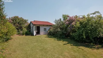 Kleines Gartenhaus umgeben von viel Grün und einer weitläufigen Rasenfläche unter blauem Himmel.