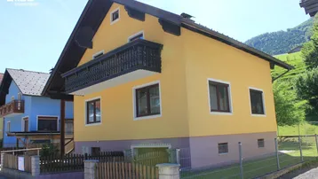 Gelbes Einfamilienhaus mit traditionellem Holzbalkon und Blick auf die umliegende Berglandschaft.