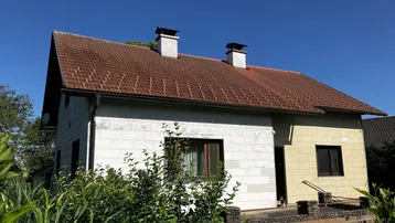 Vorderansicht des Hauses mit teilweise verputzter Fassade und Satteldach unter blauem Himmel.