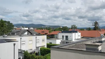 Blick über die Dächer einer Wohnsiedlung mit modernen Gebäuden und traditionellen Ziegeldächern vor bergiger Kulisse.