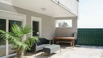 Überdachte Terrasse mit gemütlichen Sitzgelegenheiten und einem kleinen Tisch, umgeben von einer grünen Hecke.