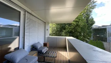 Geräumiger Balkon mit Holzboden und bequemen Sitzmöbeln, umgeben von viel Grün.
