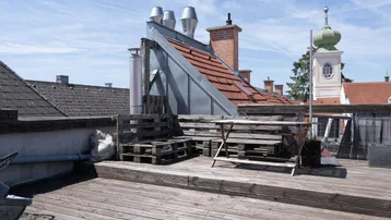 Geräumige Dachterrasse mit Holzbelag und Blick auf die umliegenden Dächer und einen Kirchturm.