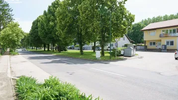 Straße mit Baumreihe, Grünstreifen und einem gelben Wohnhaus unter blauem Himmel.