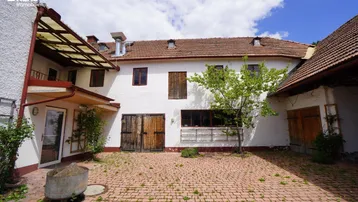 Innenhof eines alten Gebäudes mit gepflastertem Bereich, Holztüren und einem kleinen Baum in der Mitte.