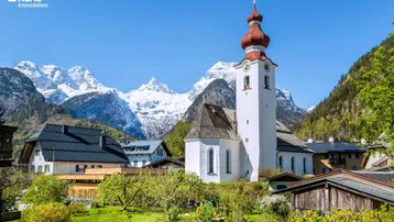 Idyllisches Dorf mit traditioneller Kirche und schneebedeckten Bergen im Hintergrund unter blauem Himmel.