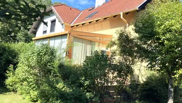 Seitenansicht des Hauses mit großem Wintergarten und üppiger grüner Bepflanzung im Garten.