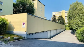 Lange Reihe von Garagenboxen mit Zufahrt und dahinterliegenden Wohngebäuden unter blauem Himmel.