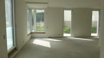 Heller, unmöblierter Wohnraum im Rohbauzustand mit großen Fenstern und Zugang zum Außenbereich.