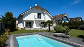 Weißes Einfamilienhaus mit privatem Pool, großer Holzterrasse und gepflegtem Garten unter blauem Himmel.