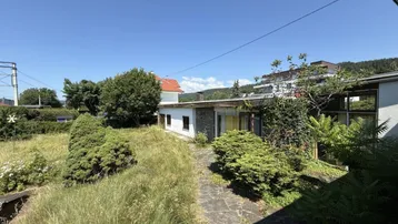 Blick auf das Haus mit verwildertem Garten und Steinweg unter blauem Himmel.