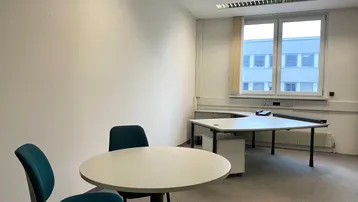 Helles Büro mit Teppichboden, einem Schreibtisch und einem kleinen Besprechungstisch am Fenster.