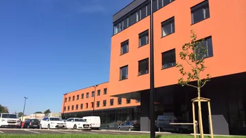 Modernes Gebäude mit oranger Fassade, großen Fenstern und Parkplätzen unter blauem Himmel.
