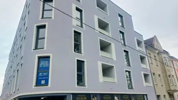 Modernes Eckgebäude mit hellgrauer Fassade, großen Fenstern und integrierten Balkonen in städtischer Umgebung.