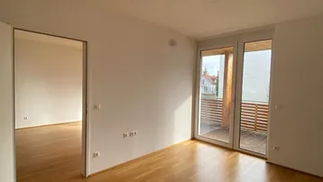 Helles, leeres Zimmer mit Parkettboden und Zugang zu einem Balkon, ideal für Wohnzwecke.