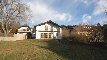 Weißes Haus mit Balkon und großem Garten auf einem Hügel unter blauem Himmel.