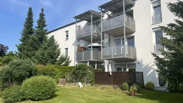 Fassade eines Mehrfamilienhauses mit mehreren Balkonen und Erdgeschoss-Terrassen, umgeben von Grünflächen.