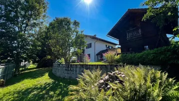 Sonniges Grundstück mit gepflegtem Garten, umgeben von Bäumen und einem traditionellen Holzhaus im Hintergrund.