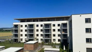 Modernes Mehrfamilienhaus mit Balkonen und Grünflächen, umgeben von Natur und Feldern.