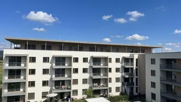 Modernes Mehrfamilienhaus mit mehreren Balkonen und Grünflächen unter blauem Himmel.