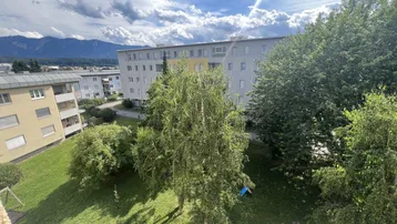 Blick auf die Wohnanlage mit grünen Bäumen und Bergen im Hintergrund unter bewölktem Himmel.