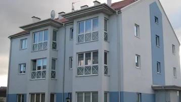 Mehrfamilienhaus mit weiß-blauer Fassade, vielen Fenstern und Satteldach unter bewölktem Himmel.