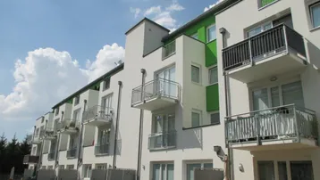 Moderne Wohnanlage mit weißer Fassade, grünen Akzenten und mehreren Balkonen sowie möblierten Terrassen im Erdgeschoss.