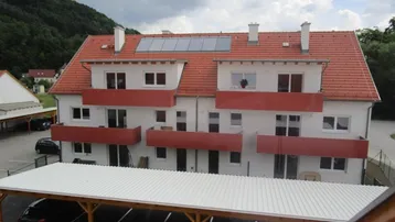 Mehrfamilienhaus mit rotem Ziegeldach, Balkonen und überdachten Parkplätzen in grüner Umgebung.