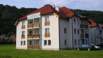 Mehrfamilienhaus mit rotem Ziegeldach und mehreren Balkonen, umgeben von Grünflächen und Hügeln.