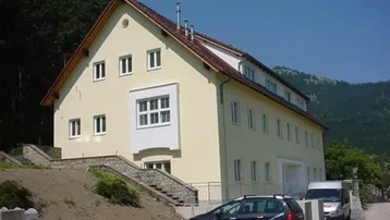 Helles Mehrfamilienhaus mit Satteldach und Bergblick, umgeben von Bäumen und Natur.