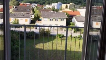 Blick von einem Balkon auf eine Wohnsiedlung mit grünen Hügeln im Hintergrund und viel Tageslicht.