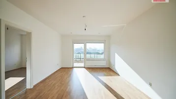 Helles, leeres Zimmer mit Holzboden und Zugang zu einem Balkon oder einer Terrasse.