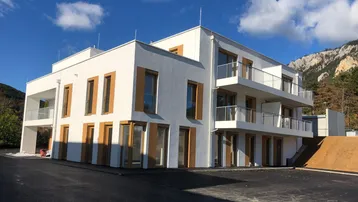 Modernes Mehrfamilienhaus mit weißer Fassade, großen Fenstern und Balkonen vor bergiger Kulisse.